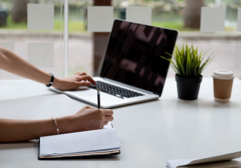 pessoa escrevendo em um caderno em cima de uma mesa com um computador do lado 
