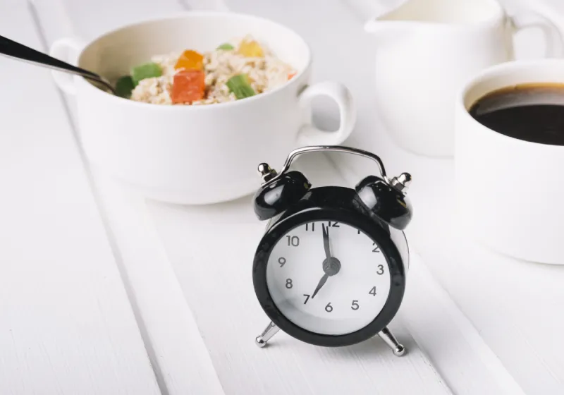 relógio despertador em cima da mesa e com xicara de café e travessa pequena com aveia e frutas dentro 