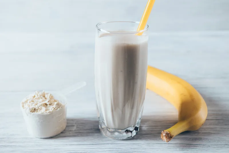 mesa com recipiente com whey, copo com vitamina e banana