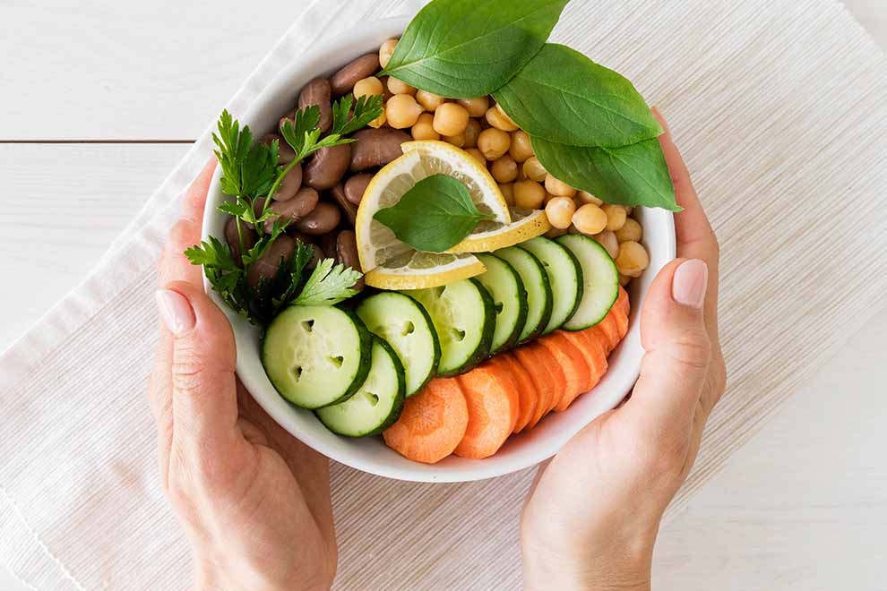 alimentação balanceada uma mão segurando um bowl de ceramica amonrrazado com salada de folhas, grãos, pepino e tomate dentro e em cima de uma mesa