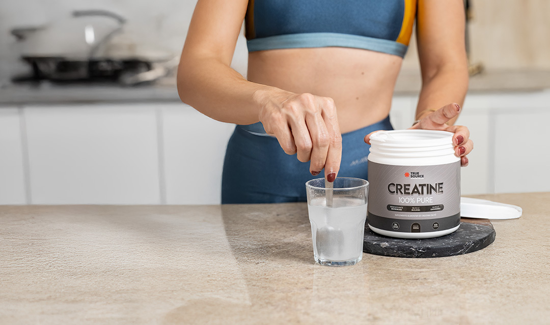 Mulher misturando um pó branco em um copo com água, ao lado de um pote de creatina da marca True Source.