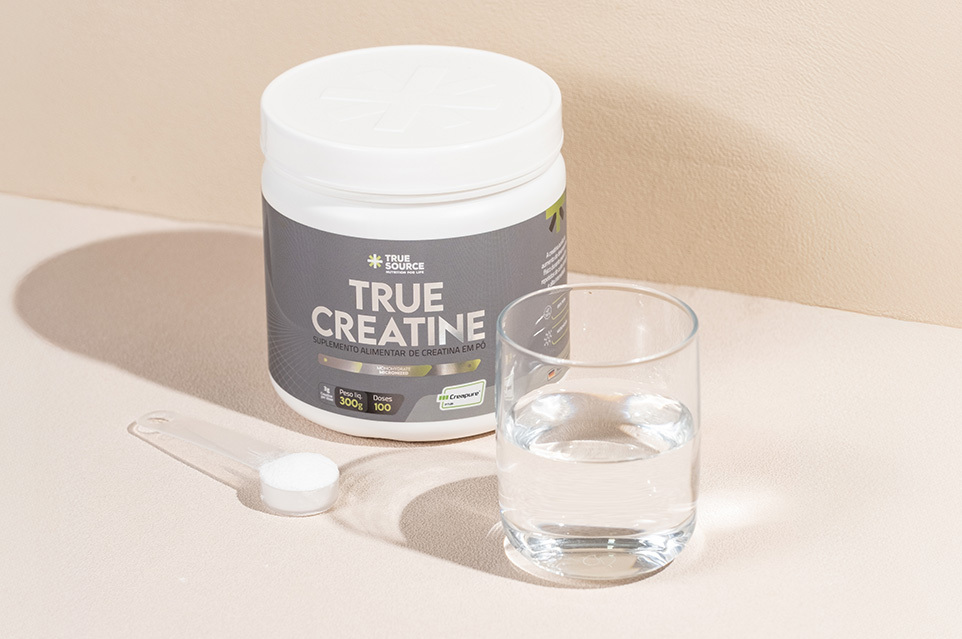 Pote de creatina True Source, um copo com água e uma colher de medida com o suplemento.