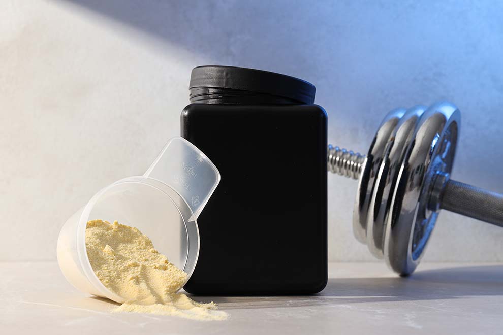 um pote grande de creatina com um copinho com a creatina em cima de uma mesa de madeira e pesos de musculação ao lado 