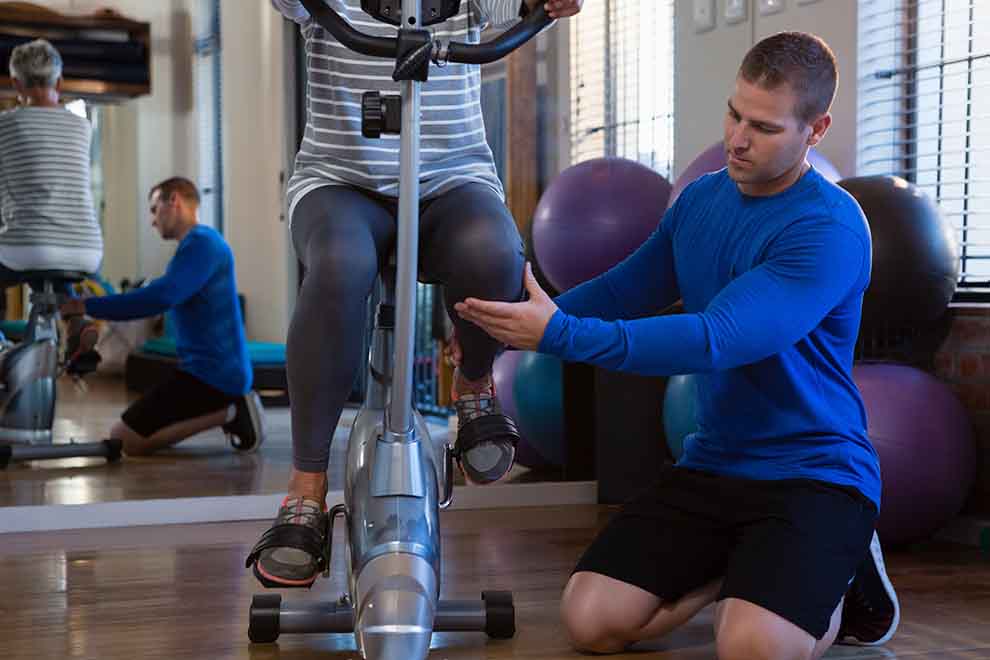 fisioterapia Treinador auxilia aluno em bicicleta ergométrica