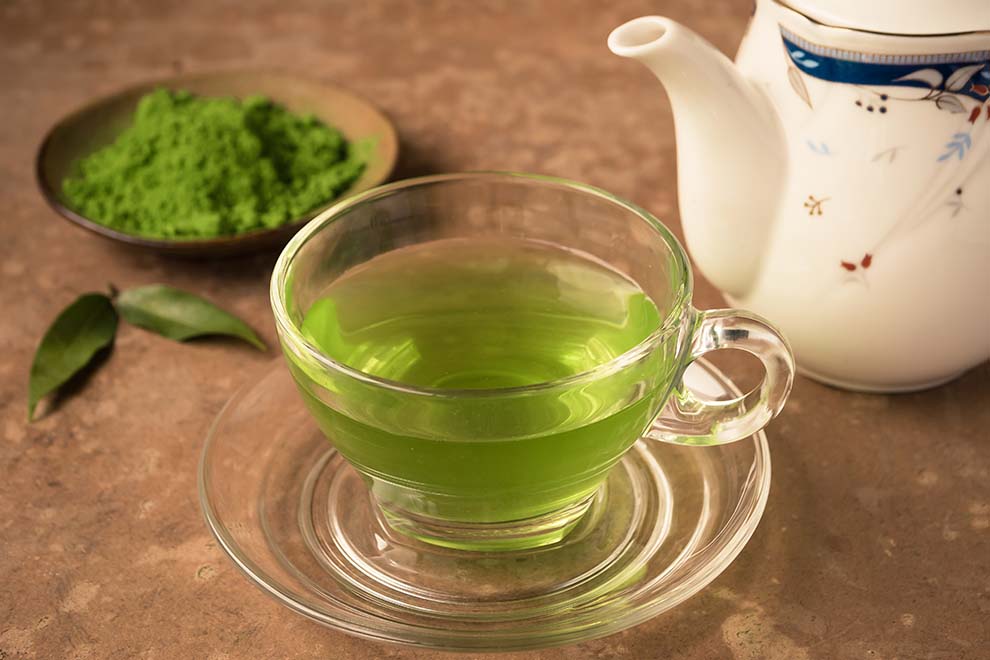 chá verde dentro de uma xicara de vidro transparente, um bule de chã de porcelana branca ao lado e um pequeno bowl com pó de chá de verde junto de um misturador de bambu tradicional japones em cima de uma mesa de madeira